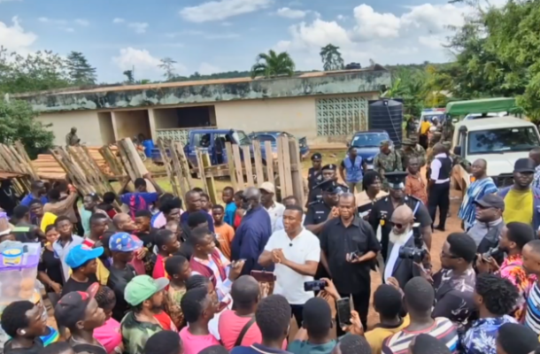 A large community gathering outdoors as a man in a white shirt speaks to a crowd, with wooden planks and vehicles in the background.