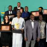 Group of award recipients posing with framed plaques on a stage during a ceremony.