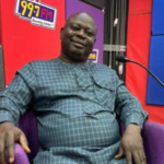 Smiling man in a blue checkered outfit sits in a purple chair in a radio studio with 99.7 FM signage nearby.