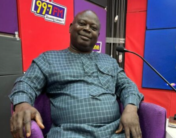 Smiling man in a blue checkered outfit sits in a purple chair in a radio studio with 99.7 FM signage nearby.