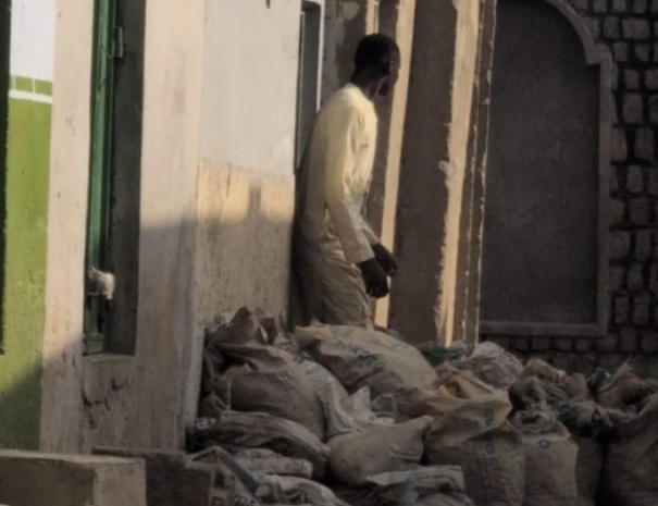 Man in light clothing stands beside a doorway on a narrow street, with many pale bags stacked on the ground.