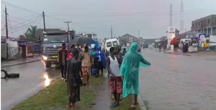 Group of people wearing rain ponchos and jackets stand on a flooded street beside moving trucks during heavy rain.