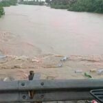 Floodwaters overflow a road with debris and plastic bottles floating; a metal guardrail is visible in the foreground.