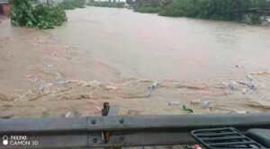 Floodwaters overflow a road with debris and plastic bottles floating; a metal guardrail is visible in the foreground.