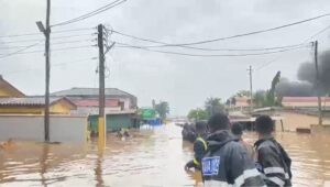 Rescue workers in reflective jackets walk through a flooded residential street with water up to mid-calf and submerged buildings nearby.