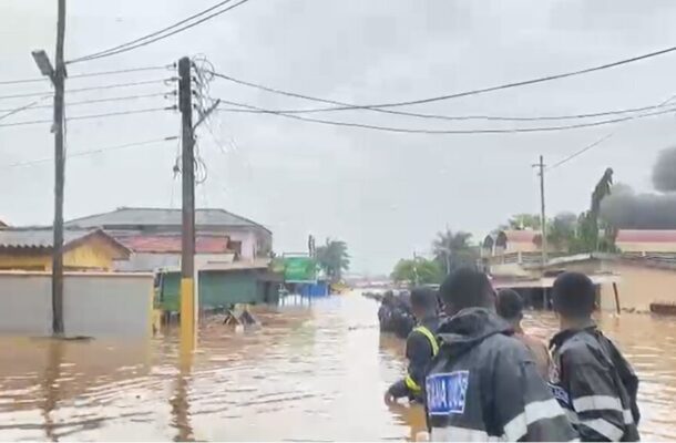 Rescue workers in reflective jackets walk through a flooded residential street with water up to mid-calf and submerged buildings nearby.