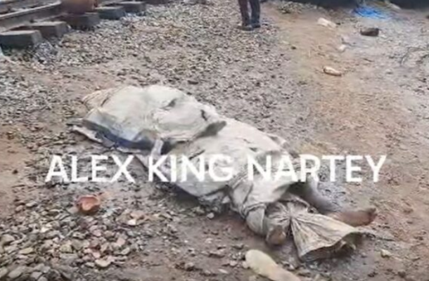 A motionless body covered with a blanket lies on rocky ground at a construction area, with a watermark reading ALEX KING NARTEY across the scene.