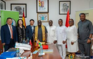Eight officials in formal and traditional attire pose for a photo in a government office, with flags, documents on the table, and portraits on the wall.