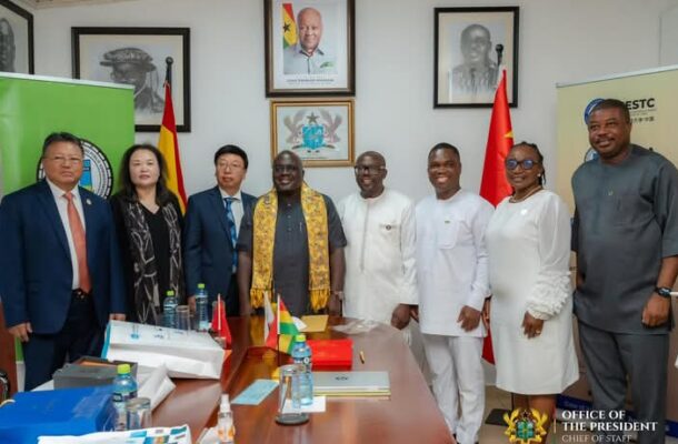 Eight officials in formal and traditional attire pose for a photo in a government office, with flags, documents on the table, and portraits on the wall.
