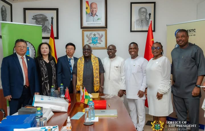 Eight officials in formal and traditional attire pose for a photo in a government office, with flags, documents on the table, and portraits on the wall.