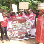 Protesters hold a large banner reading 'Say No to Abnormal Light Bills and Estimated Bills' during a demonstration; people in red cluster around the banner with signs above.