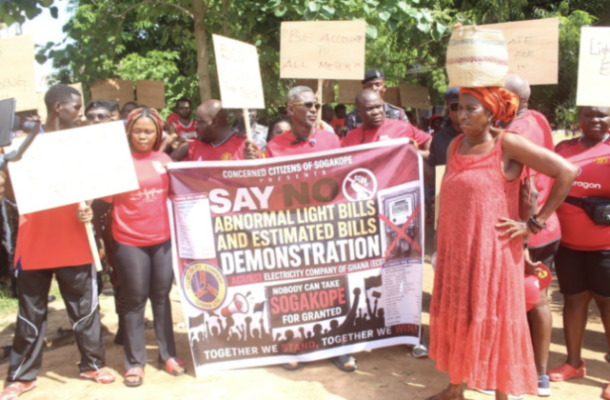 Protesters hold a large banner reading 'Say No to Abnormal Light Bills and Estimated Bills' during a demonstration; people in red cluster around the banner with signs above.