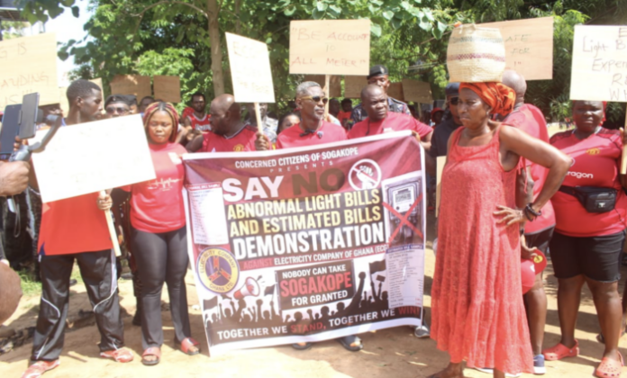 Protesters hold a large banner reading 'Say No to Abnormal Light Bills and Estimated Bills' during a demonstration; people in red cluster around the banner with signs above.