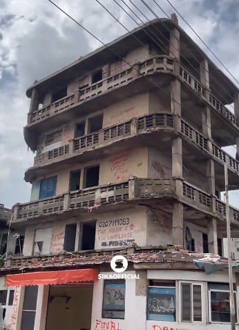 A dilapidated five-story concrete building with broken windows, graffiti on the walls, and a small street-level shop with a red awning.