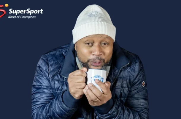 Man wearing a white knit beanie and dark jacket drinks from a white mug; SuperSport World of Champions logo visible on a dark blue background.