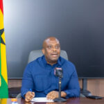Man in a blue shirt at a desk with papers and a microphone, giving a press briefing, with a Jamaican flag in the background and a crest in the corner.