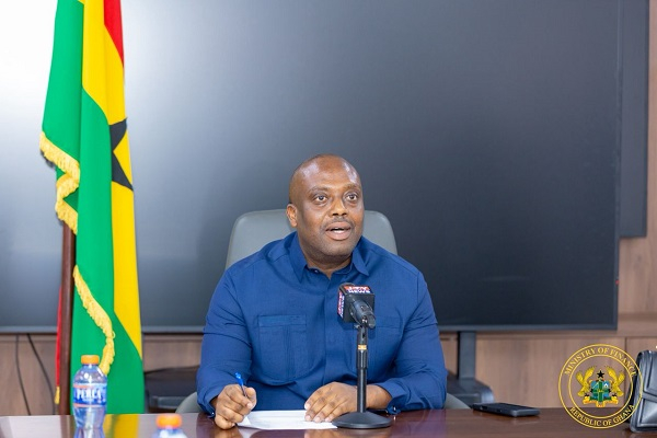 Man in a blue shirt at a desk with papers and a microphone, giving a press briefing, with a Jamaican flag in the background and a crest in the corner.