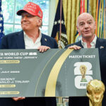 Two men in suits hold a large mock FIFA World Cup 2026 final ticket board, with an American flag and gold trophy graphic in the foreground/backgrounds evident behind them.