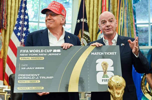 Two men in suits hold a large mock FIFA World Cup 2026 final ticket board, with an American flag and gold trophy graphic in the foreground/backgrounds evident behind them.