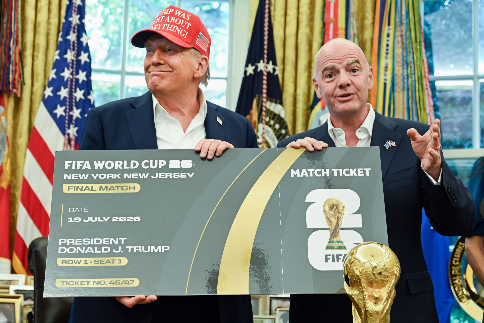 Two men in suits hold a large mock FIFA World Cup 2026 final ticket board, with an American flag and gold trophy graphic in the foreground/backgrounds evident behind them.