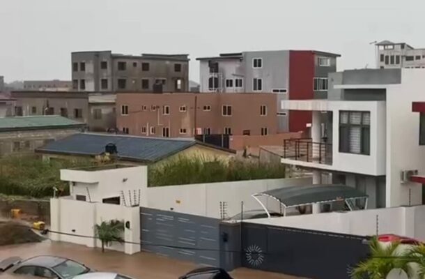 Flooded residential street with several partially submerged cars and muddy water surrounding modern houses and walls; grey, overcast sky