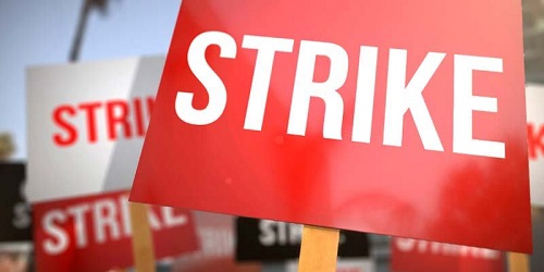 Red protest sign reading STRIKE held aloft at a labor rally, with other signs blurred in the background.