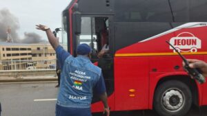 Man wearing a blue vest that reads 'ACCRA METROPOLITAN ASSEMBLY AMA MAYOR' signals to a red Jeoun bus, with smoke rising in the background.