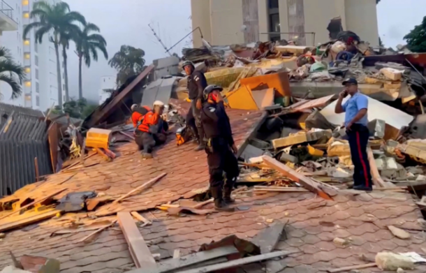 Rescue workers in orange vests search through rubble on a collapsed building roof amid urban debris.
