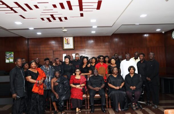 Formal group portrait of men and women in black attire, posing in a wood-paneled meeting room with a framed portrait on the wall behind them and a patterned floor.