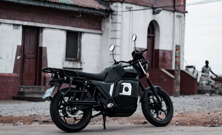 Black and gray motorcycle parked on a gravel street in front of a white building with red trim and doors visible behind it