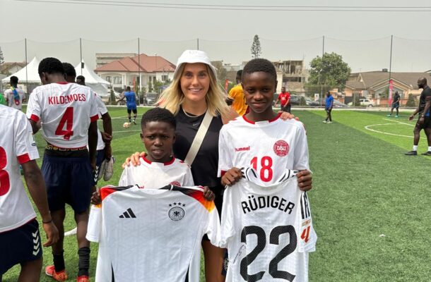 Smiling woman in a white cap stands with two young footballers on a grassy field, holding their white jerseys up front.