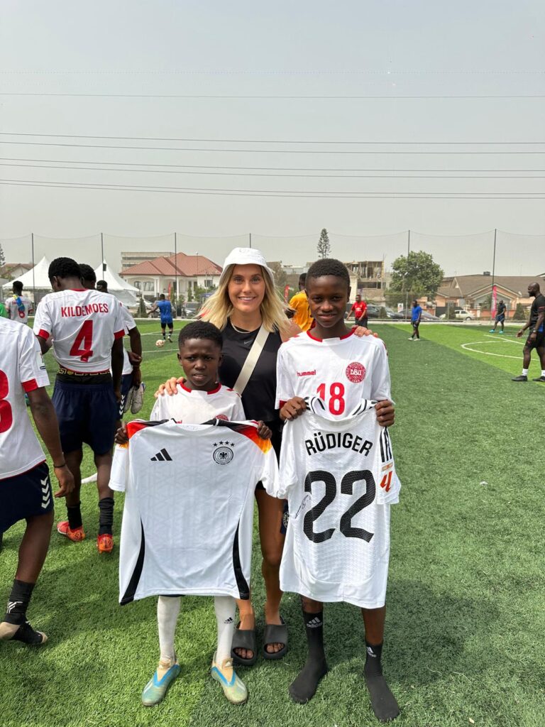 Smiling woman in a white cap stands with two young footballers on a grassy field, holding their white jerseys up front.