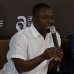 Man in a white short-sleeve shirt speaks into a microphone during a panel discussion.