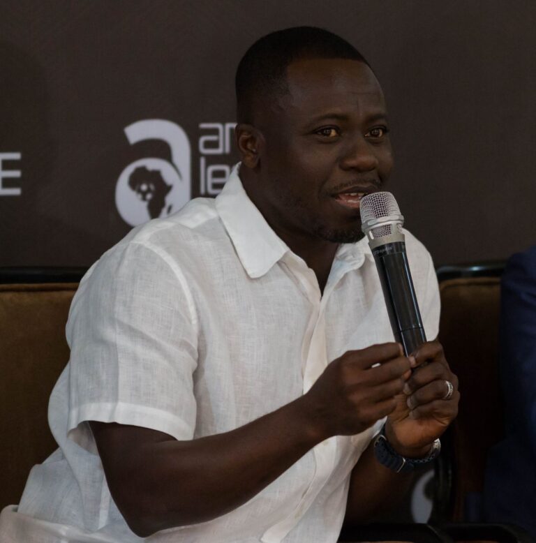 Man in a white short-sleeve shirt speaks into a microphone during a panel discussion.