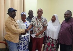 Six people stand in a room as two men shake hands and exchange envelopes, others watching nearby.