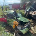 Severely damaged green car after a crash in a grassy roadside area, with a toppled truck nearby and bystanders in the background.