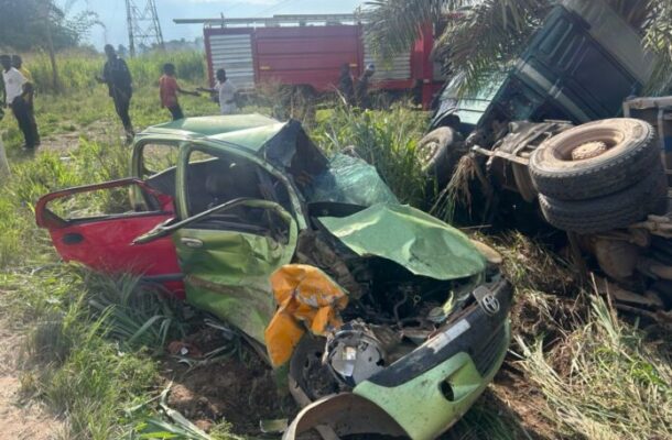 WhatsApp-Image-2026-06-13-at-221459-696x522 | Ghanaguardian.com Severely damaged green car after a crash in a grassy roadside area, with a toppled truck nearby and bystanders in the background.