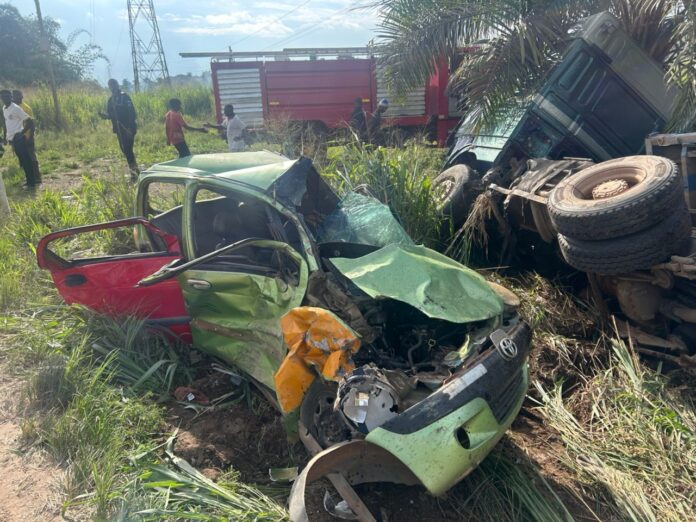 WhatsApp-Image-2026-06-13-at-221459-696x522 | Ghanaguardian.com Severely damaged green car after a crash in a grassy roadside area, with a toppled truck nearby and bystanders in the background.