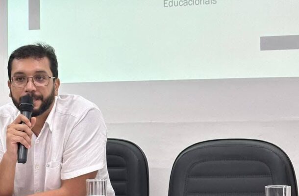 Man with glasses in a white shirt speaks into a microphone in a meeting room, with two black chairs visible behind him.