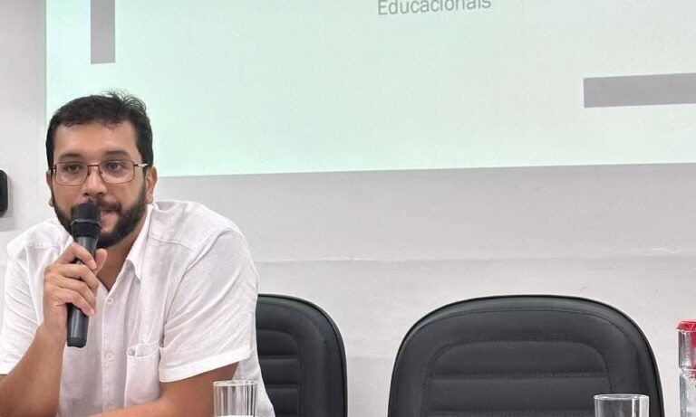 Man with glasses in a white shirt speaks into a microphone in a meeting room, with two black chairs visible behind him.