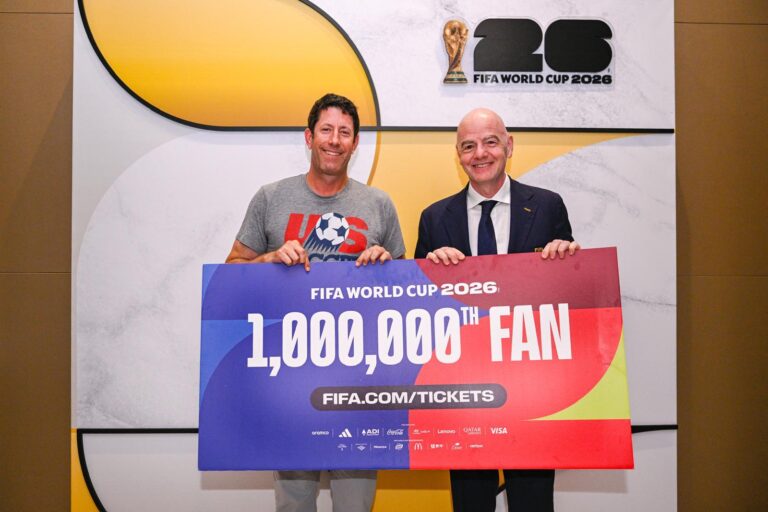Two men smiling and holding a large colorful sign reading '1,000,000th FAN' at a FIFA World Cup 2026 event, with a backdrop featuring the tournament logo behind them.