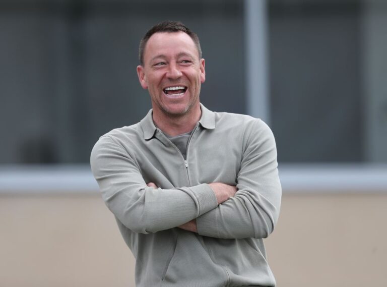 Man laughing with arms crossed outdoors, wearing a light gray quarter-zip sweater.