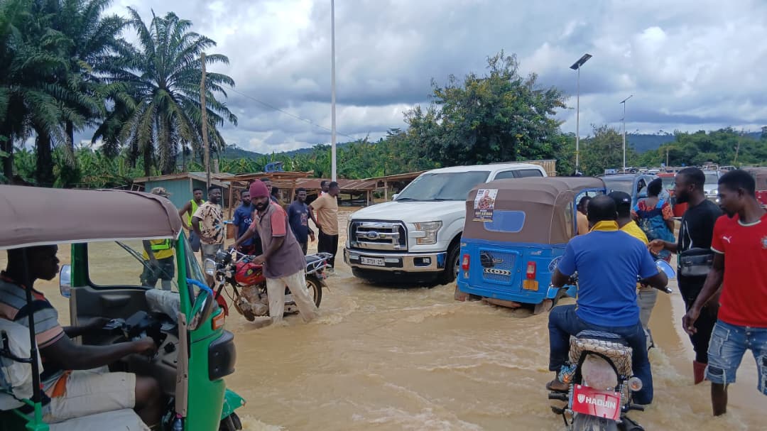 WhatsApp-Image-2026-06-18-at-33744-PM | Ghanaguardian.com Street flooded with muddy water; people on motorcycles and in tuk-tuks navigate between vehicles nearby.