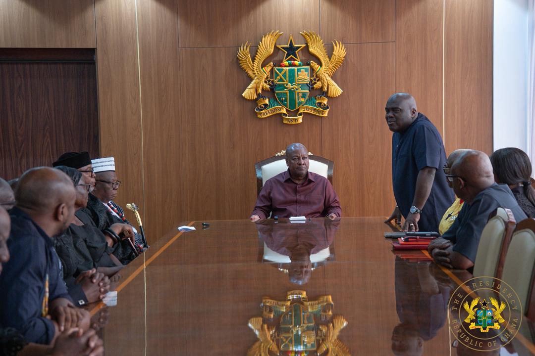 President-like figure seated at head of large wooden conference table, others around, in a formal government meeting room with a crest on the wall behind.
