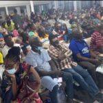 Large indoor hall filled with seated people wearing masks, waiting in rows for an event or service. Many are looking at phones or resting.