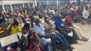 Large indoor hall filled with seated people wearing masks, waiting in rows for an event or service. Many are looking at phones or resting.