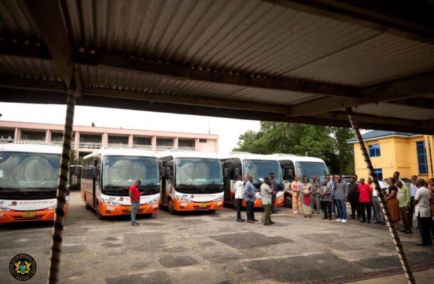 Group of people standing and chatting beside a row of orange-and-white buses at a covered bus depot.