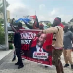 A group of protesters holding a red banner that reads 'KENNEDY MUST GO!' featuring a portrait of a man; they're outside a glass-front building in a parking lot with cars in the background.