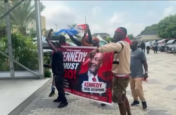 A group of protesters holding a red banner that reads 'KENNEDY MUST GO!' featuring a portrait of a man; they're outside a glass-front building in a parking lot with cars in the background.