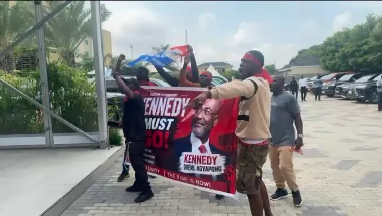 A group of protesters holding a red banner that reads 'KENNEDY MUST GO!' featuring a portrait of a man; they're outside a glass-front building in a parking lot with cars in the background.